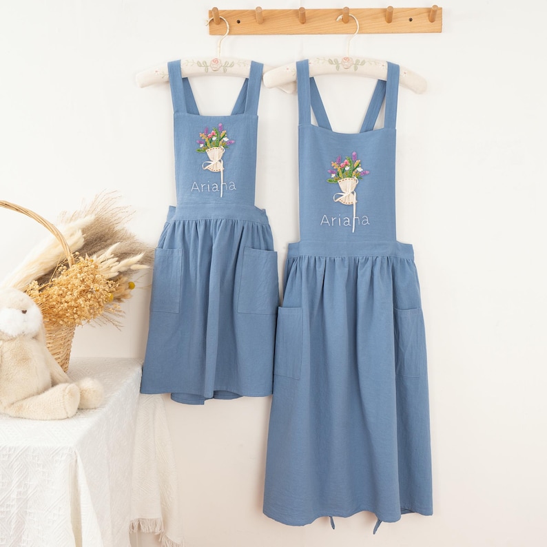 Puede incluir: Dos delantales azules colgados en una pared. Cada delantal tiene un dise&ntilde;o de bordado floral y el nombre "Ariana" bordado debajo. Los delantales tienen bolsillos y est&aacute;n colgados de perchas blancas.