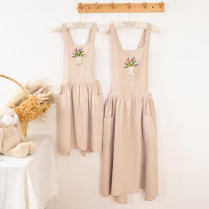 Puede incluir: Dos delantales beige colgados de un perchero de madera. Cada delantal presenta un dise&ntilde;o de bordado floral y bolsillos. Un delantal tiene el nombre "Ariana" bordado. Los delantales cuelgan de perchas blancas.