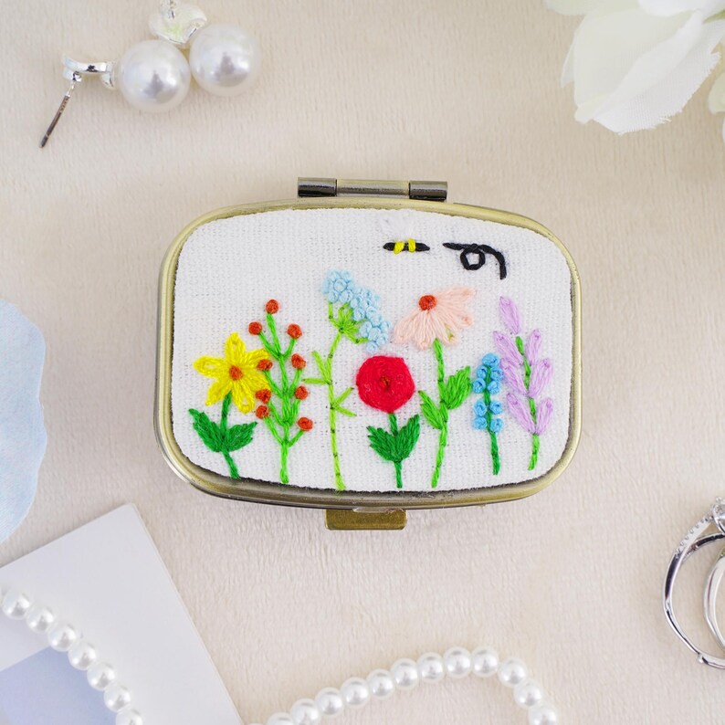 May include: A rectangular metal compact mirror featuring a white fabric cover with hand-embroidered colorful wildflowers, including yellow, red, pink, and blue blossoms with green stems, and a small black bee motif stitched near the top edge.