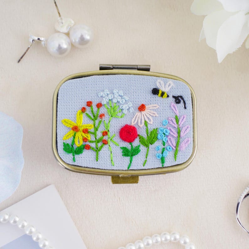 May include: An oval metal pill box featuring a light blue fabric lid embroidered with a colorful wildflower meadow and a small bee. The embroidery includes yellow, red, pink, and blue flowers with green stems. The box has a bronze-toned metal frame.