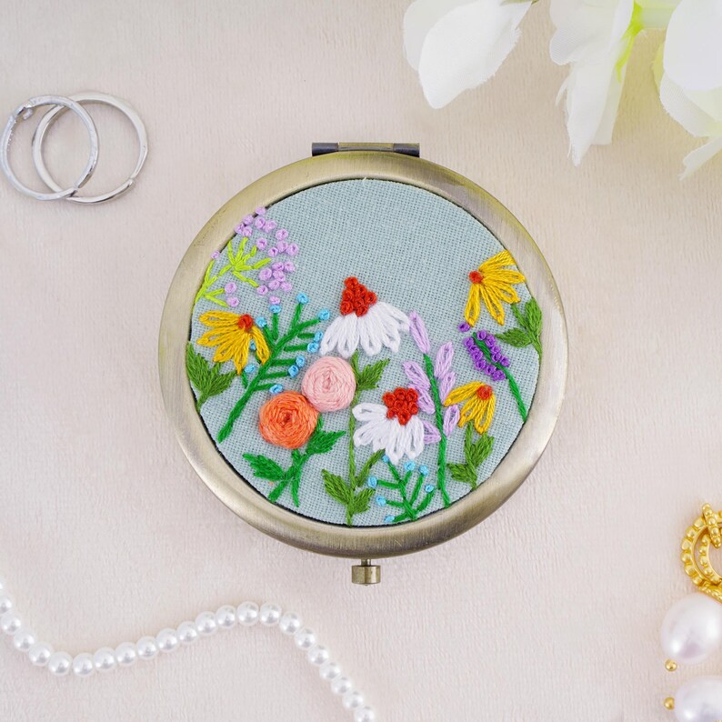 May include: A round metal compact mirror featuring a light blue fabric cover with hand-embroidered wildflowers. The floral design includes white, yellow, orange, and pink petals with green stems and leaves. The compact has a metallic bronze-toned frame.
