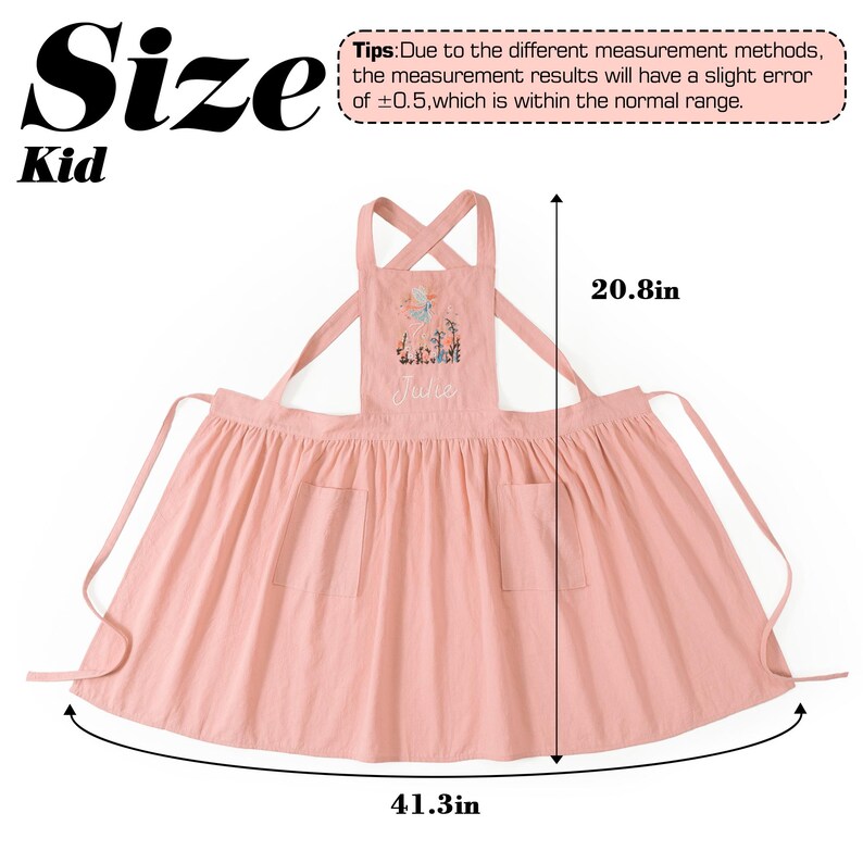 May include: A light pink child's apron featuring a floral embroidery and the name Jule on the chest. The apron has two front pockets and crisscross straps. Dimensions are 20.8 inches in height and 41.3 inches in width as indicated by arrows.