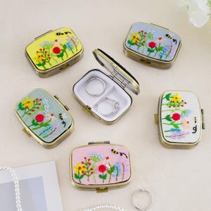 May include: A collection of rectangular metal jewelry cases with hinged lids featuring embroidered floral and bee designs in yellow, light blue, sage green, white, and pink. One case is open, revealing a white interior with two silver rings.
