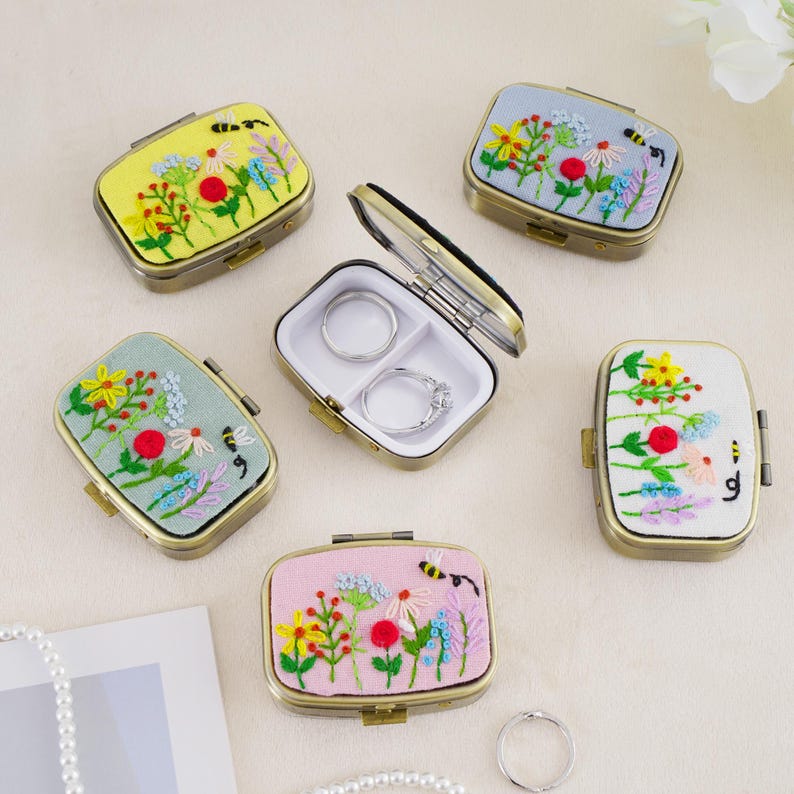 May include: A collection of rectangular metal jewelry cases with hinged lids featuring embroidered floral and bee designs in yellow, light blue, sage green, white, and pink. One case is open, revealing a white interior with two silver rings.