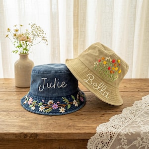 May include: A dark blue denim bucket hat embroidered with the name Julie and a floral border, and a tan bucket hat embroidered with the name Bella and a colorful wildflower bouquet. Both hats feature detailed stitching and fabric construction.