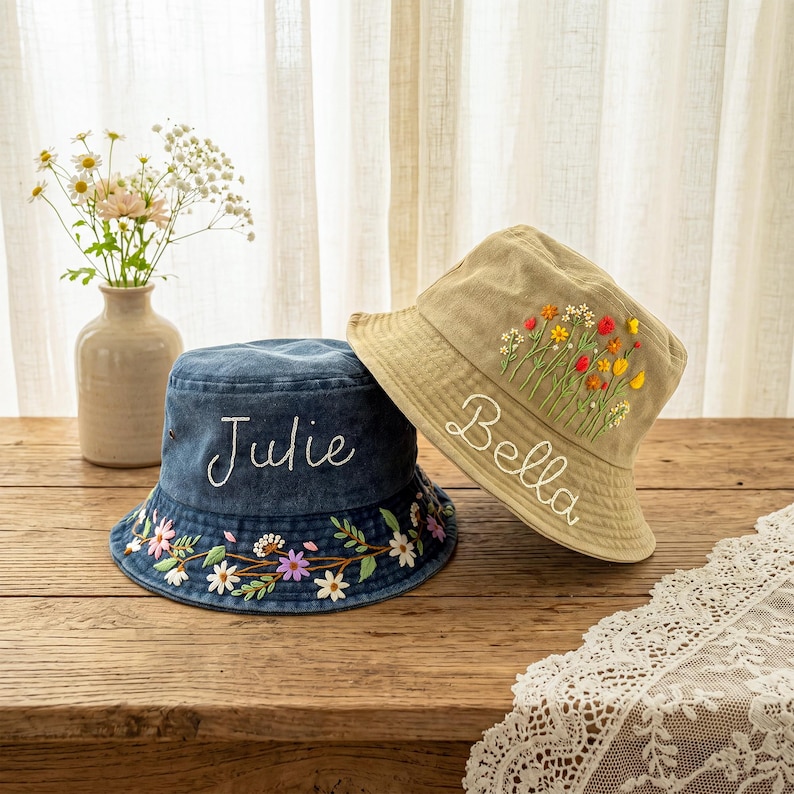 May include: A dark blue denim bucket hat embroidered with the name Julie and a floral border, and a tan bucket hat embroidered with the name Bella and a colorful wildflower bouquet. Both hats feature detailed stitching and fabric construction.