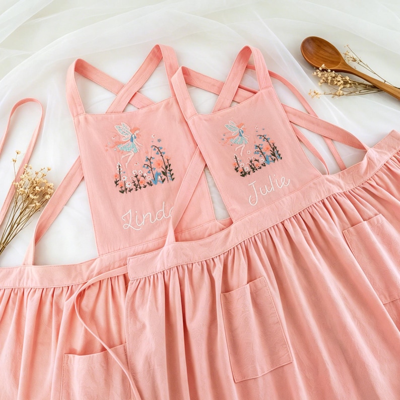 May include: Two matching dusty pink aprons with cross-back straps and front pockets. Each apron features an embroidered fairy motif surrounded by colourful flowers and the names Linda and Julie stitched in white cursive thread on the chest area.