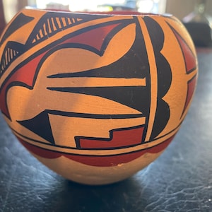 May include: A hand-painted ceramic bowl with a rounded shape. The bowl features a cream-colored base with geometric designs in black, red, and orange. The rim has a red border, and the overall design is symmetrical.