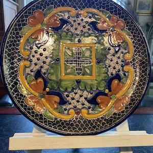 May include: A decorative ceramic plate with a dark blue scalloped border. The plate features a central square design with a cross, surrounded by floral and scroll patterns in orange, yellow, green, and blue. The plate is displayed on a white easel.