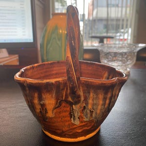 Puede incluir: Una cesta de cerámica hecha a mano con un asa retorcida. La cesta tiene un esmalte marrón y naranja con acentos goteados. El borde tiene un borde festoneado. La cesta probablemente se utiliza con fines decorativos.