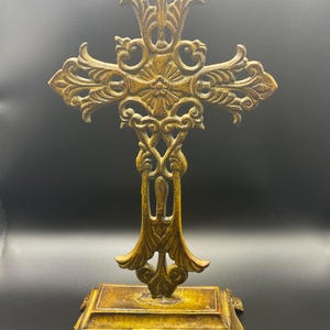 May include: Ornate, golden-colored metal cross with intricate scrollwork and a decorative base. The cross stands upright, showcasing detailed patterns and a vintage aesthetic. The base is rectangular with decorative feet.