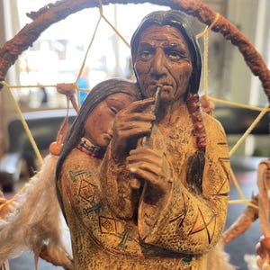 Puede incluir: Una escultura detallada de dos figuras nativas americanas, una tocando una flauta, la otra apoyando la cabeza en el hombro de la primera figura. Las figuras visten ropa tradicional, con detalles intrincados y un atrapasueños en el fondo.