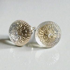 Könnte beinhalten: Zwei transparente Glasschrankknöpfe mit floralem Design. Die Knöpfe haben einen silberfarbenen Metallsockel und eine klare, abgerundete Glasoberseite mit einem detaillierten, geprägten Blumenmuster. Geeignet für Möbel oder Dekorationszwecke.