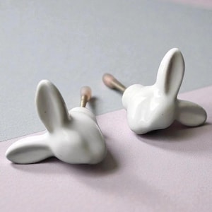 May include: Two white ceramic bunny-shaped cabinet knobs. Each knob features a bunny head with long ears and a small metal screw. The knobs are set against a soft, two-tone background of lavender and grey.