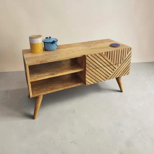 May include: A light brown wooden media console featuring two open shelves on the left and two drawers on the right with a diagonal slat pattern. The unit stands on four tapered wooden legs and holds a yellow canister and a blue metal pot.
