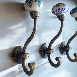 May include: A trio of wall-mounted coat hooks. Each has a bronze-toned, curved metal arm and a ceramic knob with blue floral designs on a white base. The hooks are fixed to a white wall.