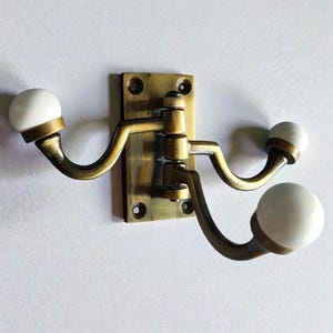 May include: A brass-toned wall hook with three curved arms, each ending in a white, spherical knob. The hook is mounted on a rectangular backplate with screw holes, suitable for hanging items. The design is vintage-inspired.