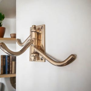 May include: A brass double hook wall mount. The hook has a rectangular backplate with two curved hooks. The hooks are mounted on a swivel, allowing them to fold flat against the wall. The finish is a polished brass.