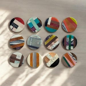 Może przedstawiać: Dwunastu okrągłych podkładek pod kubki z geometrycznymi wzorami wykonanymi z różnych rodzajów drewna i kamienia. Podkładki są w różnych kolorach, w tym brązowym, czarnym, białym, czerwonym, zielonym i niebieskim.