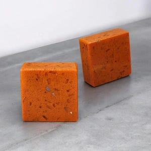 May include: Two orange-colored, cube-shaped objects with a speckled texture. The cubes are positioned on a gray surface, with one cube slightly angled. The objects have a smooth, polished appearance.
