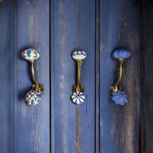 May include: Three decorative ceramic hooks with floral designs mounted on a blue wooden wall. The hooks are made of brass and have a curved shape. The ceramic designs are in shades of blue, white, and red.