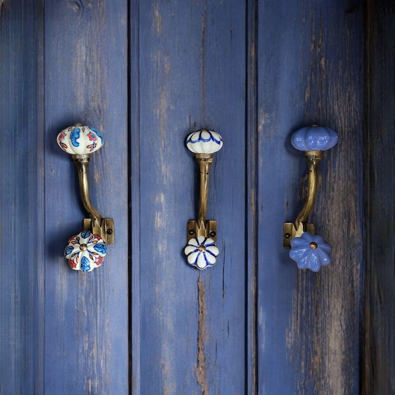 May include: Three decorative ceramic hooks with floral designs mounted on a blue wooden wall. The hooks are made of brass and have a curved shape. The ceramic designs are in shades of blue, white, and red.