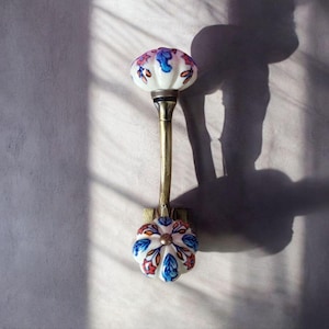 May include: A white ceramic hook with a floral design in blue, pink, and orange. The hook is attached to a brass rod and has a decorative knob at the top.