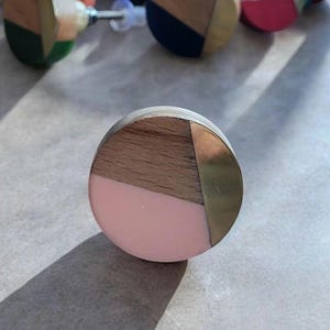 May include: A round cabinet knob with a wooden top, a pink lower section, and a brass accent. Other knobs with similar designs are visible in the background. The knob is a decorative hardware element.