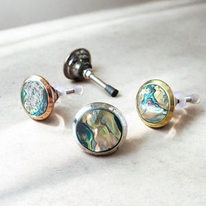 Könnte beinhalten: Vier Schmuckstücke werden gezeigt. Zwei Ohrringe mit schillernden Abalone-Muscheln sind in gold- und kupferfarbenem Metall gefasst. Ein silberfarbener Ohrring und eine kleine Metallpfeife sind ebenfalls vorhanden. Die Artikel befinden sich auf einer hellen Oberfläche.