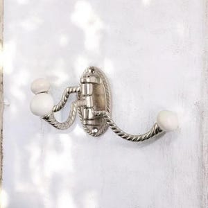 K&ouml;nnte beinhalten: Ein silberfarbener Metall-Wandhaken mit zwei gebogenen Armen und wei&szlig;en Keramikkn&ouml;pfen. Der Haken hat ein dekoratives seilartiges Design an den Armen und eine zentrale ovale Montageplatte. Das Design ist von Vintage inspiriert.