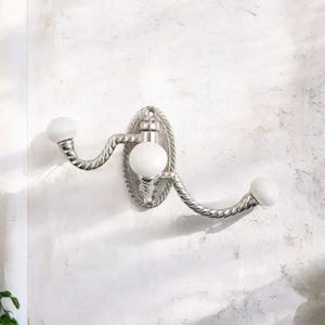 K&ouml;nnte beinhalten: Ein silberfarbener Wandhaken mit ovaler R&uuml;ckplatte und zwei geschwungenen Armen. Jeder Arm endet in einem wei&szlig;en, kugelf&ouml;rmigen Knauf. Das Design weist eine seilartige Textur an den Armen und am Rand der R&uuml;ckplatte auf.