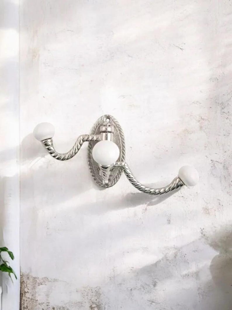 K&ouml;nnte beinhalten: Ein silberfarbener Wandhaken mit ovaler R&uuml;ckplatte und zwei geschwungenen Armen. Jeder Arm endet in einem wei&szlig;en, kugelf&ouml;rmigen Knauf. Das Design weist eine seilartige Textur an den Armen und am Rand der R&uuml;ckplatte auf.
