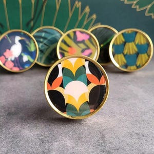 May include: Set of six round gold cabinet knobs with colourful geometric patterns. The patterns include a white bird on a blue background, green and yellow leaves, pink and yellow flowers, green and yellow stripes, and a black and white geometric pattern.
