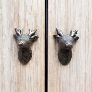 Puede incluir: Dos tiradores de armario con forma de cabeza de ciervo en color bronce, montados en puertas de armario de madera clara. Las cabezas de ciervo tienen cuernos detallados y un acabado texturizado. Las puertas están separadas por una línea vertical negra.