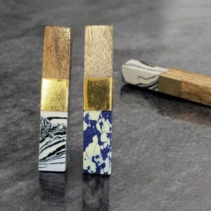 May include: Two decorative bottle stoppers with gold accents. One stopper has a wood top and a black and white marbled design. The other has a wood top and a blue and white speckled design. A third stopper is partially visible in the background.