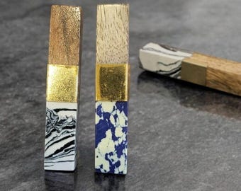 May include: Two decorative bottle stoppers with gold accents. One stopper has a wood top and a black and white marbled design. The other has a wood top and a blue and white speckled design. A third stopper is partially visible in the background.