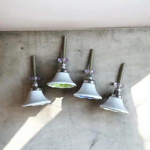 K&ouml;nnte beinhalten: Vier wei&szlig;e Keramik-Schrankkn&ouml;pfe mit Blumendesigns. Jeder Knopf hat einen silbernen Metallschaft und eine durchsichtige violette Kunststoffscheibe.