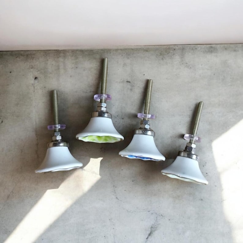 K&ouml;nnte beinhalten: Vier wei&szlig;e Keramik-Schrankkn&ouml;pfe mit Blumendesigns. Jeder Knopf hat einen silbernen Metallschaft und eine durchsichtige violette Kunststoffscheibe.