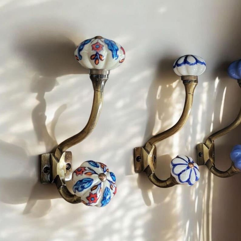 May include: Three decorative wall hooks with ceramic knobs. The knobs are painted with floral patterns in blue, white, and orange. The hooks are made of brass and have a curved shape.