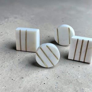 Könnte beinhalten: Vier dekorative Schrankknöpfe in verschiedenen Formen: zwei Quadrate und zwei Kreise. Jeder Knopf ist weiß mit goldfarbenen Streifen. Die Knöpfe sind auf einer neutralfarbenen Oberfläche angeordnet.