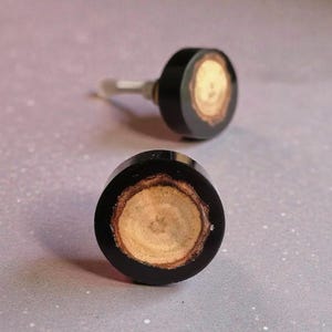 May include: Two cabinet knobs with a black surround and a light brown wood-grain centre. One knob has a silver stem. The knobs are circular in shape and are likely for furniture.