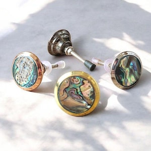May include: An assortment of decorative cabinet knobs and a bottle stopper. The knobs feature iridescent abalone shell insets with gold, copper, and silver-toned metal rims. The bottle stopper has a silver-toned metal top and a flexible, silver-toned metal stem.