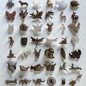 Op de afbeelding: Een verzameling kleine, decoratieve metalen dierenfiguren in verschillende tinten bruin, brons en wit. De collectie omvat bijen, honden, vogels, herten, vissen, egels, vlinders, uilen, zeepaardjes, libellen en meer. Dit zijn waarschijnlijk kastknoppen of decoratieve accenten.