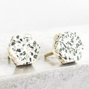 Puede incluir: Dos pomos hexagonales para armario con base blanca y un patrón moteado de manchas negras y verdes.