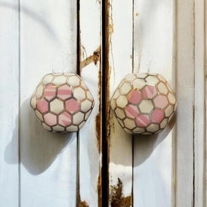 Może przedstawiać: Dwa dekoracyjne gałki do szafek. Każda gałka ma geometryczny wzór z kremowym tłem, różowymi akcentami i złotymi konturami. Zamontowane na białej, postarzonej drewnianej powierzchni.