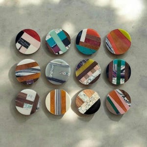 May include: Twelve round coasters with geometric patterns made from colourful wood and stone. Each coaster features a unique combination of materials and colours, including turquoise, brown, red, green, white, and black.