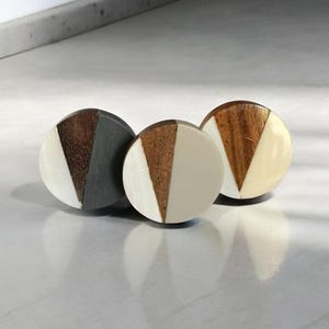 May include: Three round wooden and white knobs with a geometric design. The knobs are in a row on a white surface.