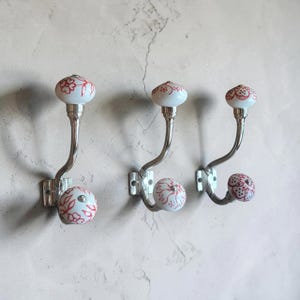 May include: Three wall-mounted coat hooks made of polished silver-tone metal. Each hook features two white ceramic knobs decorated with intricate red floral patterns. The hooks are arranged in a row, each with a curved arm and a square base plate.