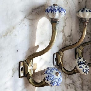 May include: Antique brass hooks with blue and white ceramic knobs. The hooks are mounted on a white marble wall.