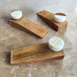 May include: Three wooden door stops with white circular tops. One top has a gold stripe, another has a gold half-circle, and the third has a black half-circle. The door stops are made of light brown wood and are arranged on a marble surface.